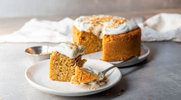 Karotten-Nusskuchen mit Frischkäse-Topping, Im Vordergrund liegt ein Stück Kuchen mit einer Gabel auf einem weißen Keramikteller, im Hintergrund steht der restliche Kuchen auf einem Teller, ganz hinten sieht man ein weißes Tischtuch