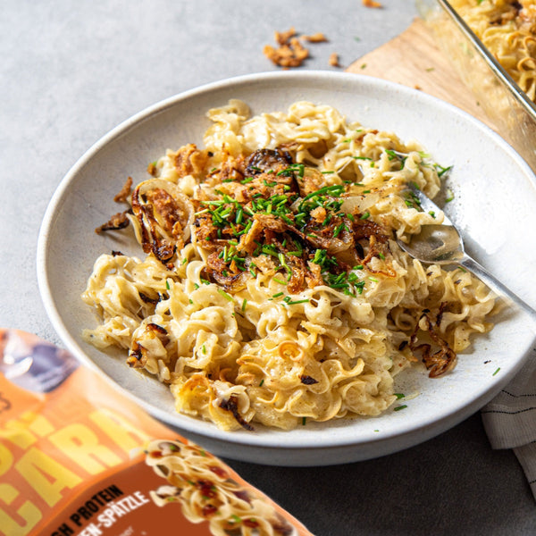 Auf einem weißen Teller liegt eine Portion Käsespätzle mit vielen Röstzwiebeln und etwas Schnittlauch darauf. Daneben liegt eine orangene Verpackung von STEINER´s Low Carb High Protein Locken Spätzle, die verwendet wurden.