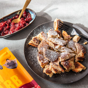 Zubereiteter STEINER´s Low Carb Kaiserschmarrn mit Puder-Erythit bestäubt. Der Kaiserschmarrn ist auf einem schwarzen Teller serviert. Links daneben ist ein Schälchen mit Beerenkompott und eine Packung Kaiserschmarrn.