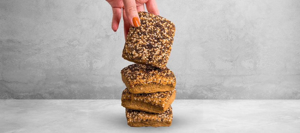 Dreit STEINER´s Low Carb High Proten Saatenbrötchen sind übereinander gestapelt auf einer grauen Fläche. Ein viertes Brötchen wird gerade senkrecht von einer Hand auf den Stapel gehalten. So sind die Saaten zum Betrachter gerichtet.