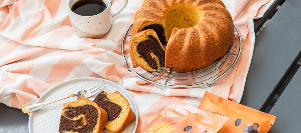 Marmorkuchen in Ringform steht angeschnitten auf einem orange- weißen Tischtuch. Daneben liegen zwei orangene STEINER´s Low Carb Kuchen Backmischungen, eine Tasse Kaffee und ein weißer Kuchenteller mit zwei Sttück Marmorkuchen darauf und einer Gabel