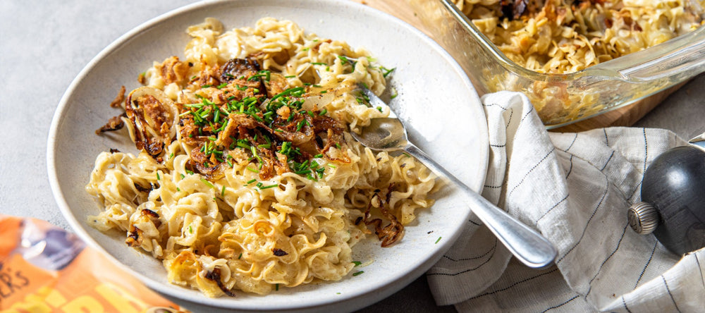 Auf einem weißen Teller liegt eine Portion Käsespätzle mit vielen Röstzwiebeln und etwas Schnittlauch darauf. Daneben liegt eine orangene Verpackung von STEINER´s Low Carb High Protein Locken Spätzle, die verwendet wurden.