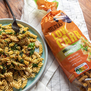 STEINER´s Low Carb High Protein Fusilli in vegan. Links ist ein blauer Teller mit den zubereiteten Fusilli. Die Soße besteht aus grünem Pesto, frischen Kräutern und STEINER´s Pasta Gewürz. Rechts danaben liegt eine volle Packung der Fusilli.