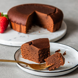 ein Stück STEINER´s Schokokuchen auf einem weißen kleinem Teller im Vordergrund. Auf einer goldenen kleinen Gabel steckt ein Stück Schokokuchen.  Im Hintergrund ist der ganze Kuchen angeschnitten zu sehen und daneben liegen zwei Erdbeeren.