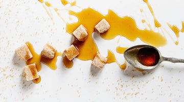 Sirup ist auf einem weißen Tisch verteilt. In dem Sirup liegt ein Teelöffel und mehrere Zuckerwürfel.