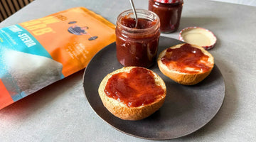 Ein Glas Erdbeermarmelade steht neben einem belegten Brötchen und einer Packung STEINER´s Low Carb Erythrit + Stevia.