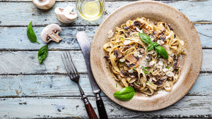 STEINER´s Tagliatelle mit gebratenem Ingwer-Gemüse. Angerichtet auf einem braunem Teller. Neben dem Teller befinden sich aufgeschnittene weiße Champignons und vintage-Besteck.