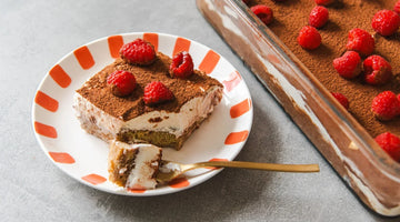 Ein Stück cremiges Tiramisu auf einem weißen Teller mit orangenen Streifen am Rand. Das Tiramisu ist mit Kakao und Himbeeren getoppt. Rechts ist das Tiramisu in der Backform.