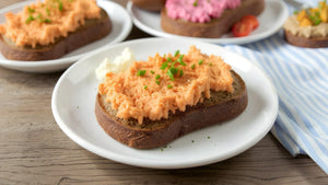 Eine Schreibe STEINER´s Brot mit einer Liptauer-Creme liegt auf einem wißen Teller. Der Teller staht auf einem blau weiß gestreiften Tuch. Zu sehen ist ein Holztisch, ein Laib STEINER´s Brot.