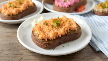 Eine Schreibe STEINER´s Brot mit einer Liptauer-Creme liegt auf einem wißen Teller. Der Teller staht auf einem blau weiß gestreiften Tuch. Zu sehen ist ein Holztisch, ein Laib STEINER´s Brot.