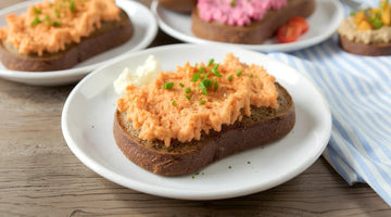 Eine Schreibe STEINER´s Brot mit einer Liptauer-Creme liegt auf einem wißen Teller. Der Teller staht auf einem blau weiß gestreiften Tuch. Zu sehen ist ein Holztisch, ein Laib STEINER´s Brot.
