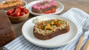 Eine Schreibe STEINER´s Brot mit einer Creme aus Pfidderlingen liegt auf einem wißen Teller. Der Teller staht auf einem blau weiß gestreiften Tuch. Zu sehen ist ein Holztisch, ein Laib STEINER´s Brot.