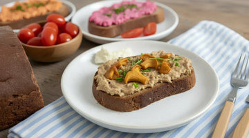 Eine Schreibe STEINER´s Brot mit einer Creme aus Pfidderlingen liegt auf einem wißen Teller. Der Teller staht auf einem blau weiß gestreiften Tuch. Zu sehen ist ein Holztisch, ein Laib STEINER´s Brot.