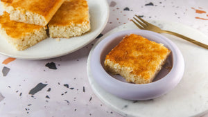 Quadratische Stücke eines Blechs selbstgebackener Kokosschnitten. Ein Stück liegt auf einem kleinen Teller in lila. Der untergrund ist Terrazzo.