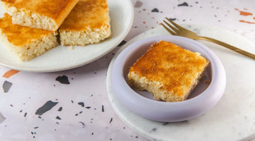 Quadratische Stücke eines Blechs selbstgebackener Kokosschnitten. Ein Stück liegt auf einem kleinen Teller in lila. Der untergrund ist Terrazzo.