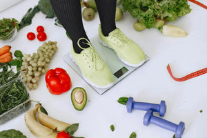 Eine Frau steht auf einer Waage. Sie trägt eine schwaze Leggings und hellgrüne Turnschuhe. Neben ihr befinden sich kleine Hanteln und frisches obst und Gemüse auf dem Boden.