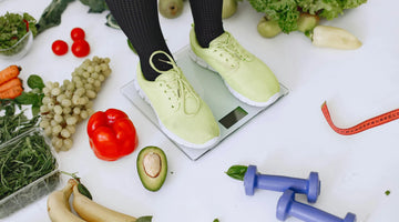 Eine Frau steht auf einer Waage. Sie trägt eine schwaze Leggings und hellgrüne Turnschuhe. Neben ihr befinden sich kleine Hanteln und frisches obst und Gemüse auf dem Boden.