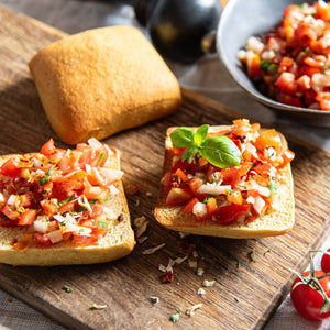 Auf einem Holzbrett liegt ein ganzes STEINER´s Low Carb High Protein Brötchen und zwei Brötchenhälften belegt mit Bruschetta. Dahinter ist eine Schale mit Bruschetta zu sehen.