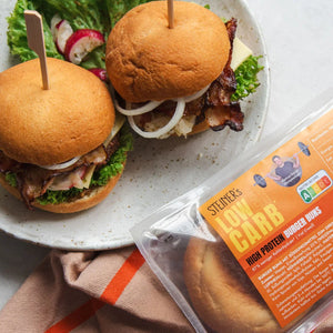 Auf einem Holzbrett liegt ein ganzes STEINER´s Low Carb High Protein Brötchen und zwei Brötchenhälften belegt mit Bruschetta. Dahinter ist eine Schale mit Bruschetta zu sehen.