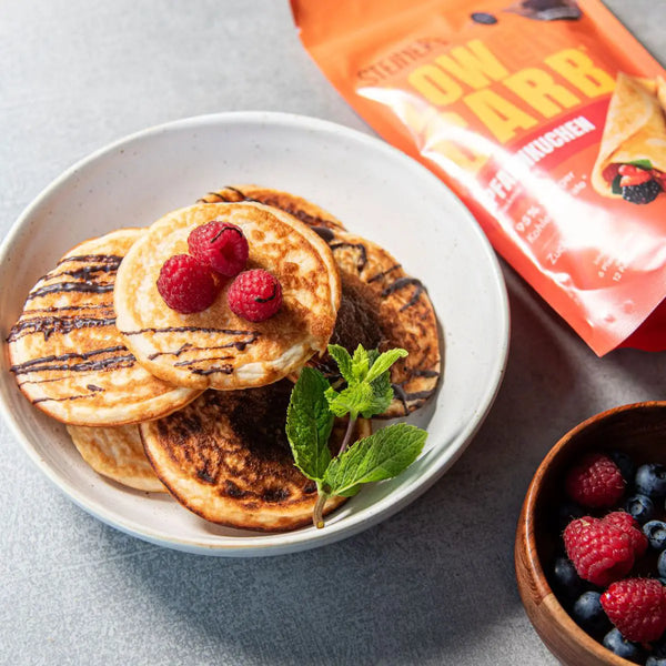 Auf einem weißen Teller liegen sechs Stück Pancakes, gebacken aus unserer STEINERé Low Carb Pfannkuchen Backmischung. Neben den Pancakes befindet sich die Pfannkuchenverpackung und eine Holzschale mit Himbeeren und Heidelbeeren.