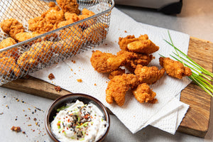 6 Chicken nuggets liegen auf einem Holzbrett, weitere in einem Fritierkorb. Alle wurden mit STEINER´s Low Carb High Protein Semmelbrösel paniert und ausgebacken. Daneben steht eine Schale mit einem weißen Jogurt Dip mit Gewürzen.