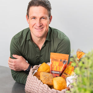 Olypiasieger Matthias Steiner im grünen Hemd lehnt mit seinen verschränkten Armen auf einer Tischplatte. er lächelt. Vor ihm steht ein Brotkörchen gefüllt mit unterschiedlichen STEINER´s Low Carb Produkten