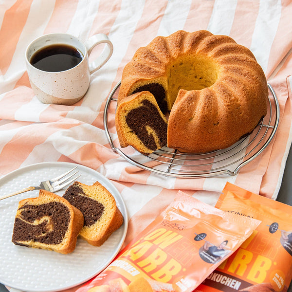 Marmorkuchen in Ringform steht angeschnitten auf einem orange- weißen Tischtuch. Daneben liegen zwei orangene STEINER´s Low Carb Kuchen Backmischungen, eine Tasse Kaffee und ein weißer Kuchenteller mit zwei Sttück Marmorkuchen darauf und einer Gabel
