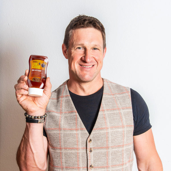 Matthias Steiner lächelt in die Kamera und hält eine Flasche STEINER´s Low Carb BBQ Ketchup in seiner rechten Hand.
