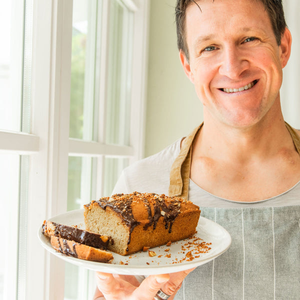 Matthias Steiner steht vor einer Fensterfront und trägt eine graue Schürze. In seiner Hand hält er einen weißen Teller mit einem STEINER´s Nusskuchen in Kastenform. Der Kuchen ist mit flüssiger Schokolade und gehackten Nüssen getoppt.