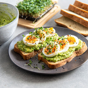Zwei STEINER´s Brotscheiben, belegt mit einer Avocadocreme, geschnittenem Ei und frischer Kresse. Die Brotscheiben liegen auf einem dunkelgrauem Teller. Im Hintergrund ist geschnittenes Brot und Kresse.