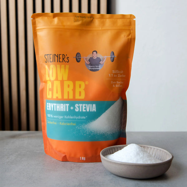 Eine Packung STEINER´s Erythrit-Stevia. Die Packung ist Orange und Blau. In der Packung Erythrit-Stevia sind 1kg. Die Packung steht vor einer Akustikpaneele und steht auf einer grauen Arbeitsplatte aus Stein. Vor der Packung ist ein graues Schälchen, gefüllt mit Erythrit-Stevia.