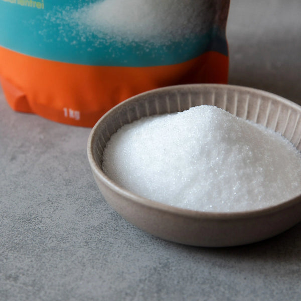 Eine Packung STEINER´s Erythrit-Stevia. Die Packung ist Orange und Blau. In der Packung Erythrit-Stevia sind 1kg. Die Packung steht auf einer grauen Arbeitsplatte aus Stein. Vor der Packung ist ein graues Schälchen, gefüllt mit Erythrit-Stevia.