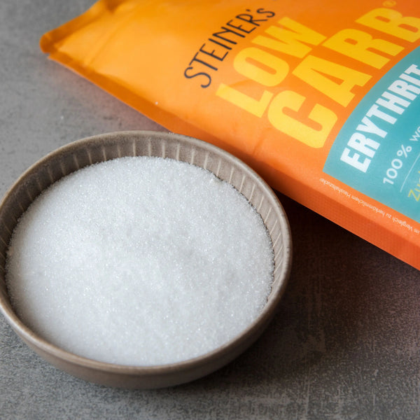 Eine Packung STEINER´s Erythrit-Stevia. Die Packung ist Orange und Blau. In der Packung Erythrit-Stevia sind 1kg. Die Packung steht auf einer grauen Arbeitsplatte aus Stein. Vor der Packung ist ein graues Schälchen, gefüllt mit Erythrit-Stevia.