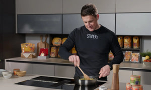 Matthias Steiner steht in einer Küche und schwenkt eine Pfanne mit Pasta.