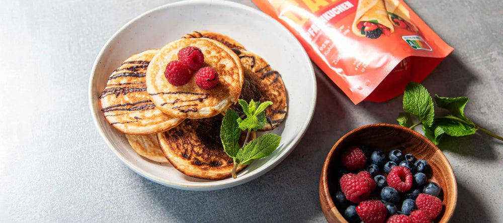 Auf einem weißen Teller liegen sechs Stück Pancakes, gebacken aus unserer STEINERé Low Carb Pfannkuchen Backmischung. Neben den Pancakes befindet sich die Pfannkuchenverpackung und eine Holzschale mit Himbeeren und Heidelbeeren.