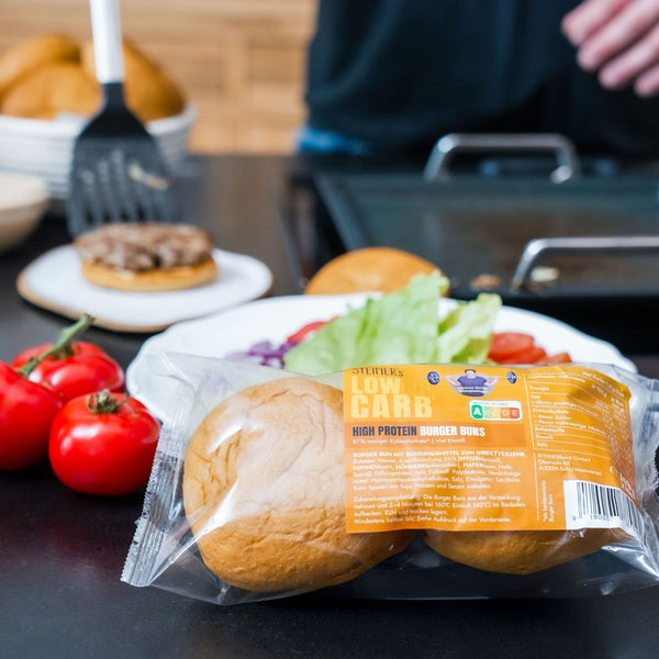 Zwei Stück STEINER´s Low Carb High Protein Burger Buns in ihrer Kunststoffverpackungen liegen auf einer Küchenarbeitsplatte. Dahinter wird ein saftiger Börger belegt.